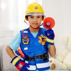 Junge in Feuerwehrweste, Helm mit Logo. Er hält ein Megafon und trägt blaue Handschuhe. Kuscheltier im Hintergrund.