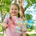 Ein lachendes Kind macht Seifenblasen mit einem pinken Blasstab im Park, Bäume im Hintergrund.