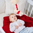 Keine sichtbaren Texte. Ein Baby liegt wach in einem weißen Gitterbett, schaut auf einen hängenden Plüschbären.