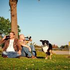 Text auf gelbem Schild: "Geringste Höhe nächster Ziel". Zwei Männer sitzen im Gras, trinken Bier. Ein Hund läuft vorbei.