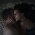 Ein Paar liegt engumschlungen im Bett, sie schauen sich zärtlich an. Dunkler Raum, ruhige, intime Atmosphäre.