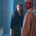 Kein Text erkennbar. Eine Frau in Schwarz schaut besorgt, ein Mann mit hellem Gewand blickt seitlich. Blaues Licht im Hintergrund.