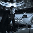 Zwei Personen in futuristischer Umgebung. Der vordere Mann trägt einen dunklen Anzug, im Hintergrund Kontrollpaneele.