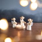 Drei Holzengel mit Instrumenten und einer Kerze auf einem Holztisch, umgeben von unscharfen Lichtern im Hintergrund.