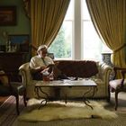 Ein älterer Mann liest auf einem beigen Sofa. Der Raum hat grüne Wände, große Fenster mit gelben Vorhängen und antike Möbel.