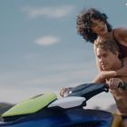 Eine Frau macht ein Selfie auf einem Jetski, während sie hinter einem Mann sitzt. Beide tragen Badebekleidung.