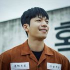 Ein Mann lächelt in brauner Uniform mit den Zahlen "2학15" und "2017" auf Schildern. Hintergrund mit unscharfer Schrift.