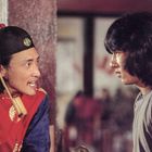 Zwei Männer interagieren. Der linke trägt traditionelle Kleidung mit rotem Gewand und Hut, der rechte hat langes, dunkles Haar.