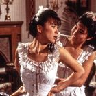 Zwei Frauen in Korsetts in einem eleganten Raum; eine hilft der anderen, ihr Korsett zu schnüren.