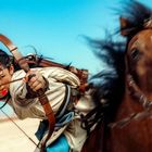 Eine Person auf einem Pferd zielt mit Pfeil und Bogen, windgepeitschtes Haar, Wüste und blauer Himmel im Hintergrund.