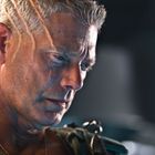 Ein Mann mit grauem, kurzem Haar, Narben auf der rechten Gesichtshälfte, schaut nachdenklich nach unten.