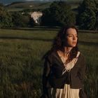 Keine sichtbaren Texte. Eine Frau steht in einem weiten grünen Feld, im Hintergrund Bäume und ein großes Gebäude.