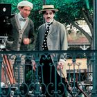 Text: "CHAS. CHAPLIN" auf einem Stuhl. Zwei Männer in altmodischer Kleidung, einer mit Schnurrbart, vor grünen Bäumen, Film-Set.