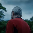 Ein Mann mit grauen Haaren in rotem Hemd steht vor einem bewölkten Himmel und Bäumen im Hintergrund.