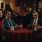 Vier Männer sitzen um einen Tisch in einem Raum mit Bar. Ein Schild zeigt "Casino". Sie unterhalten sich lebhaft.