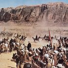Keine sichtbaren Texte. Viele Reiter in Wüstenkleidung auf Pferden, staubige Umgebung, beeindruckende Felsformationen im Hintergrund.