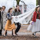 Ein Hochzeitspaar spaziert im Freien, begleitet von Freunden. Die Braut trägt ein langes Schleierkleid, der Bräutigam einen Zylinder.