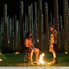 Zwei Männer, einer sitzt auf einem Stuhl, der andere steht. Ein Feuer brennt, umgeben von Totenköpfen und hohen Holzsäulen.