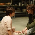 Zwei Menschen sitzen auf dem Boden einer Küche, intensiv im Gespräch. Im Hintergrund ist ungenutztes Kücheninventar erkennbar.