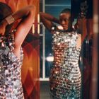 Eine Frau mit Glitzerkleid betrachtet sich in einem Spiegel, umgeben von geometrischen Mustern.