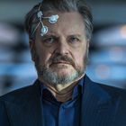 Ein älterer Mann mit grauem Haar und Bart trägt ein Gerät mit Elektroden an der Stirn. Hintergrund ist unscharf und blau.