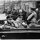 Auf einem Platzwagen sitzen Männer in Uniformen mit NS-Symbolen, Menschenmenge im Hintergrund.