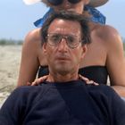 Eine Person sitzt am Strand, trägt eine Brille und sieht besorgt aus. Hinter ihr steht jemand in Sommerkleidung.