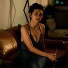 Kein Text vorhanden. Eine Frau in einem dunklen Kleid sitzt mit einem Glas Wein auf einem Ledersofa in einem gemütlichen Raum.