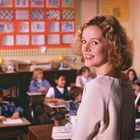 Eine Lehrerin in einem Klassenzimmer lächelt. Im Hintergrund Kinder an Schreibtischen, mit bunten Kunstwerken an der Wand.