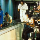 Eine Frau in blauer Uniform mit Namensschild wird auf einem Filmset von mehreren Kameraleuten begleitet.