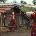Zwei Personen in bunter, traditioneller Kleidung. Eine posiert vor einer alten Hütte, die andere fotografiert sie.