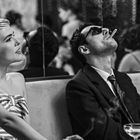 Eine Frau in gestreiftem Kleid sitzt neben einem Mann im Anzug, der eine Zigarette raucht. Beide schauen in dieselbe Richtung.