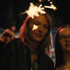 Zwei Menschen halten Wunderkerzen in der Nacht, deren Licht ihre Gesichter beleuchtet. Im Hintergrund sind Gebäude erkennbar.