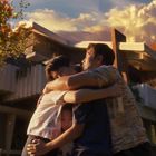Drei Personen umarmen sich vor einem modernen Haus bei Sonnenuntergang, umgeben von Bäumen und goldenem Licht.