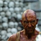 Eine Person mit dunklen Kriegsbemalungen und ernstem Blick. Hintergrund zeigt eine unscharfe Wand aus Schädeln.