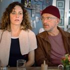 Zwei Menschen sitzen in einem Café. Eine Frau mit lockigem Haar schaut nach vorn, ein Mann mit Brille und Mütze schaut zu ihr.