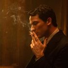 Ich kann den Text im Bild nicht identifizieren. Ein Mann in Anzug raucht nachdenklich, Rauch wirbelt in einem warm beleuchteten Raum.