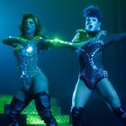 Zwei Performer in glitzernden Kostümen und Stiefeln auf einer Bühne mit grünem und blauem Licht.