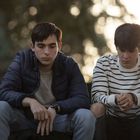 Zwei junge Männer sitzen nachdenklich nebeneinander. Einer trägt eine blaue Jacke, der andere ein gestreiftes Shirt.