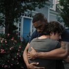 Zwei Personen umarmen sich vor einem Haus mit blauer Tür und blühenden Rosenbüschen, umgeben von Bäumen.