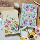 Kein Text vorhanden. Zwei bestickte Notizbücher mit Blumenmotiven, eins beige und eins blau, liegen auf einem Holztisch.