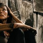 Eine Frau mit schmutzigem Gesicht sitzt nachdenklich vor kunstvollen, dunklen Zeichnungen an der Wand.