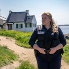 "Police Nationale" steht auf der Uniform einer Polizistin, die auf einem Küstenweg neben Häusern steht.