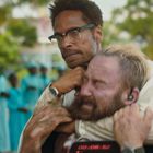 Ein Mann mit Brille hält einen anderen Mann in einem Würgegriff. Im Hintergrund stehen Menschen in türkisfarbener Kleidung.