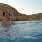 Ein Mann schwimmt im Wasser vor einer felsigen Küste. Sein Gesichtsausdruck zeigt Überraschung oder Freude.