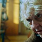 Eine ältere Frau mit lockigem, blondem Haar schaut ernst. Im Hintergrund ein unscharfer Raum mit einer Person in schwarzer Kleidung.