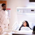 Ein "Evacuation Plan"-Schild an der Wand. Ein Mann in hellem Trainingsanzug steht neben einer Frau im Krankenhausbett.