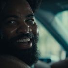 Es sind keine Texte im Bild. Ein Mann mit Bart lächelt während er in einem Auto sitzt und nach hinten schaut.