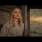 Keine erkennbaren Texte. Eine Frau mit langen, blonden Haaren steht vor einem rustikalen Gebäude und hat Tränen in den Augen.
