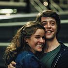 Ein lächelndes Paar umarmt sich, beide tragen lässige Kleidung, der Hintergrund ist unscharf mit Lichtern.